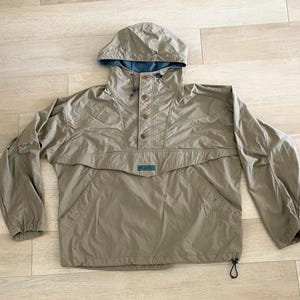 May include: A tan anorak jacket with a hood and a blue interior lining. The jacket has a button-up front, a front pocket, and drawstrings at the hem. The sleeves are long, and the jacket appears to be made of a lightweight, water-resistant material.