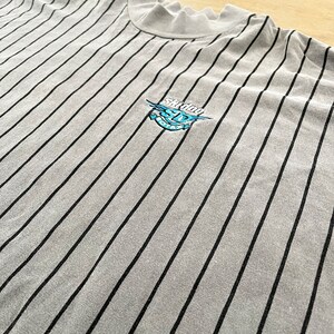 Może przedstawiać: Szara, w paski, koszula z długim rękawem z haftowanym logo w kolorze czarnym i niebieskim, na kt&oacute;rym widnieje napis "Skidder" i rysunek kresk&oacute;wkowego potwora.