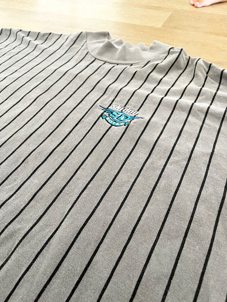 Może przedstawiać: Szara, w paski, koszula z długim rękawem z haftowanym logo w kolorze czarnym i niebieskim, na kt&oacute;rym widnieje napis "Skidder" i rysunek kresk&oacute;wkowego potwora.