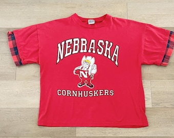 May include: Red t-shirt with a white "Nebraska Cornhuskers" logo featuring a cartoon mascot. The shirt has plaid fabric on the sleeves.