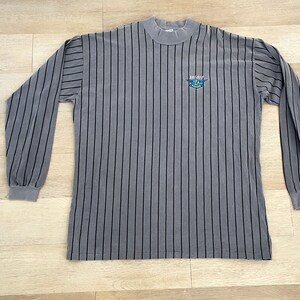 Może przedstawiać: Szara w paski koszula z długim rękawem z czarnym logo z napisem "Air City" i grafiką samolotu.