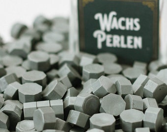 May include: A collection of gray wax seal beads scattered around a clear glass jar. The jar has a label that reads "Wachs Perlen" in white lettering on a dark green background. A wooden cork is visible in the background.