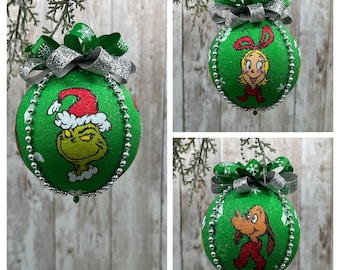 May include: Green Christmas ornaments featuring characters from a holiday story. Each ornament has a green base, silver bead trim, and a decorative bow. The characters include a grumpy green figure in a Santa hat, a blonde girl with a red bow, and a brown dog in a red scarf.