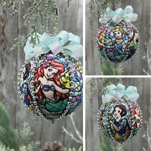 May include: A spherical Christmas ornament featuring a stained-glass style print of various animated characters. The ornament is decorated with silver beaded trim along the seams and topped with a light blue ribbon bow with iridescent glitter.