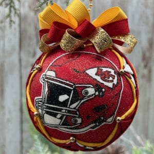 May include: A red Christmas ornament featuring a football helmet design. The ornament has a yellow and red bow at the top and is trimmed with gold accents. The ornament is designed with the team's logo.