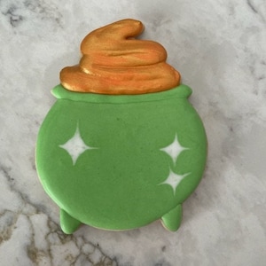 May include: Green cauldron cookie with a gold swirl on top and white glitter accents.
