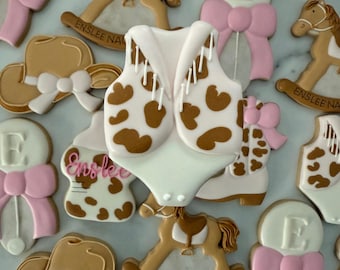 May include: A collection of decorated sugar cookies with a cowgirl theme. The cookies feature a rocking horse, a cowboy hat, a rattle, a vest, and the name "Enslee Naomi Rae".