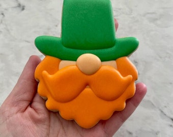 Könnte beinhalten: Ein grüner und orangefarbener Lebkuchen, der wie ein Kobold aussieht. Der Keks hat einen grünen Hut, einen orangefarbenen Bart und Schnurrbart und eine kleine orangefarbene Nase.