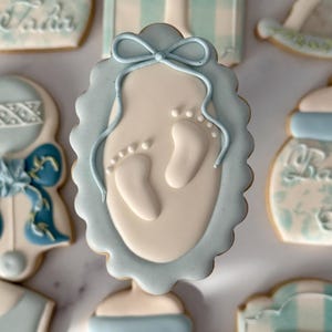 May include: An assortment of baby-themed cookies, including one with a blue oval frame and baby footprints. Other cookies feature a rocking horse, a baby bottle, and the letter "T". The cookies are decorated with light blue and white icing.