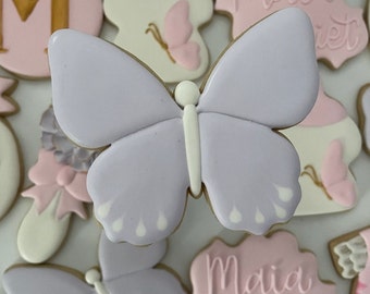 May include: A purple butterfly cookie with white accents. The cookie is decorated with a white line down the center and white dots around the edges.