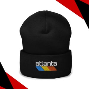 May include: A black beanie with the word "Atlanta" embroidered in white, above a horizontal stripe of blue, yellow, and orange. The hat is made of a soft, knitted material.