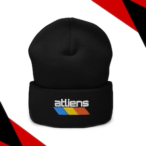 May include: Black beanie with the word "atliens" embroidered in white, above a horizontal stripe of blue, yellow, and orange. The beanie is made of a soft material and is suitable for cold weather.