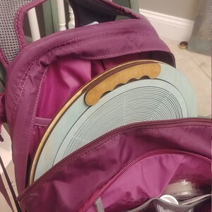 May include: A purple backpack with a zippered pocket and a round wooden disc with a handle inside. The disc is light blue with a pattern of concentric circles.