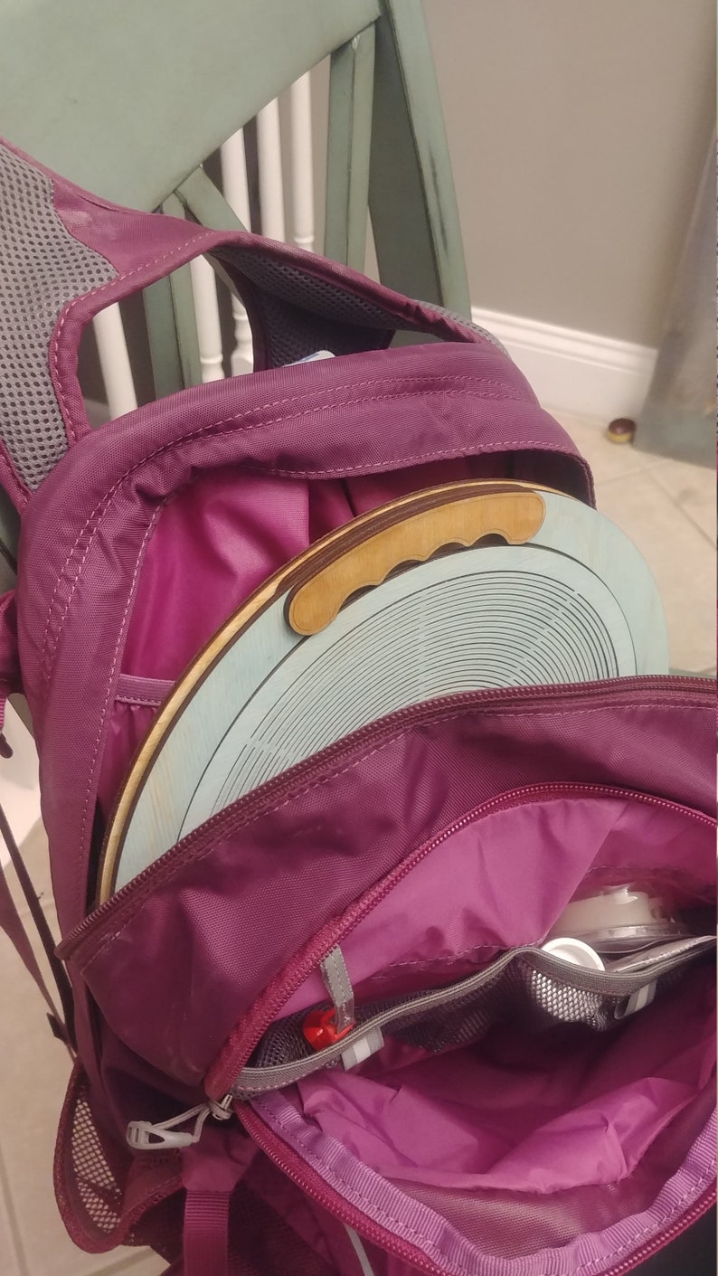May include: A purple backpack with a zippered pocket and a round wooden disc with a handle inside. The disc is light blue with a pattern of concentric circles.