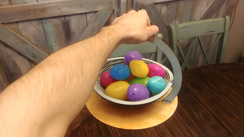 May include: A wooden basket with a handle, filled with colorful plastic Easter eggs. The basket is sitting on a wooden table.