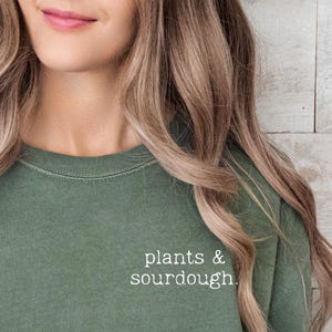 Può includere: Una donna indossa una maglietta verde con la scritta "plants & sourdough" stampata sul davanti. Indossa jeans blu e ha i capelli sciolti.