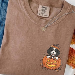 Puede incluir: Una camiseta marrón de manga corta con un gráfico de un perro blanco y negro sentado dentro de una calabaza naranja tallada. La camiseta tiene una etiqueta Comfort Colors en el cuello, se combina con vaqueros azules y está rodeada de calabazas de tela.