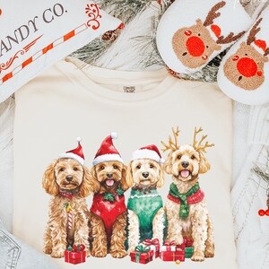Peut inclure: Un t-shirt crème avec quatre chiens vêtus d'habits de Noël. Les chiens portent des bonnets de Père Noël, des bois de rennes et des pulls festifs. Un oreiller avec des graphiques de cannes en sucre et le texte "KRINGLE CANDY CO." est également visible.