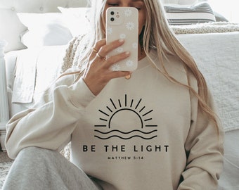 May include: A beige sweatshirt with a black graphic of a sun rising over water and the text "BE THE LIGHT" with "MATTHEW 5:14" underneath.