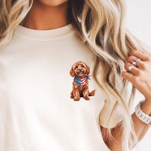 May include: Cream-colored t-shirt featuring a watercolor illustration of a brown dog wearing a bandana with an American flag design. The dog is sitting, and the shirt has a classic crew neck and short sleeves. The shirt is paired with blue jeans.