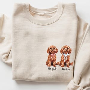 May include: A cream-coloured sweatshirt with a watercolour illustration of two brown dogs, each wearing a bow. The names "Hazel" and "Duke" are written below the dogs in a script font. The sweatshirt has long sleeves.
