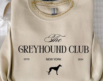 May include: Cream-colored sweatshirt with "The Greyhound Club" text and a greyhound silhouette. Includes "ESTD NEW YORK 2024" text. Paired with light-wash denim jeans, a gold chain necklace, and white high-top sneakers.