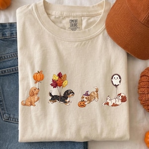 Puede incluir: Una camiseta color crema con un gráfico de cuatro perros con elementos de otoño y Halloween, incluyendo un globo de calabaza, hojas de otoño, un sombrero de bruja y un globo de fantasma, junto a unos vaqueros azules y una gorra color óxido.