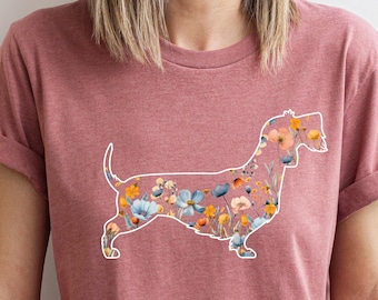 May include: A dusty rose t-shirt with a dachshund outline filled with a floral design in blue, orange, and yellow. The top has short sleeves and a round neck.