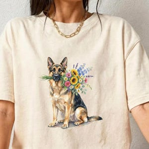 Peut inclure: T-shirt cr&egrave;me avec une illustration &agrave; l'aquarelle d'un berger allemand tenant un bouquet de fleurs color&eacute;es. Le chien est repr&eacute;sent&eacute; assis. Le t-shirt a des manches courtes et une coupe d&eacute;contract&eacute;e.