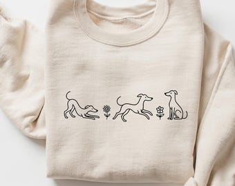 May include: Beige sweatshirt with a crew neck and long sleeves. The front features a black line drawing of three dogs in different poses, with small flower illustrations between them. The sweatshirt is made of a soft material.