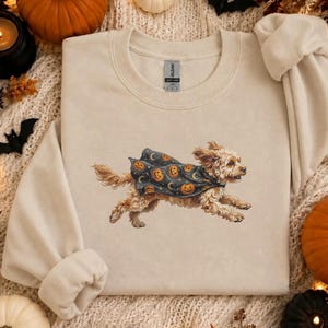 Puede incluir: Una sudadera de cuello redondo color crema con un gráfico de un perro marrón claro corriendo, vestido con una capa oscura decorada con calabazas de Halloween naranjas y lunas crecientes. La prenda tiene mangas largas y cuello acanalado.