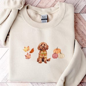May include: Cream-coloured sweatshirt with a watercolour illustration of a brown dog wearing a scarf, with autumn leaves and pumpkins. The design is centered on the front of the crewneck sweatshirt. The sleeves are rolled up.