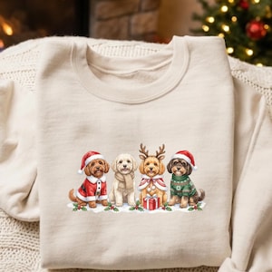 May include: A beige crewneck sweatshirt featuring a graphic print of four dogs dressed in holiday attire. The dogs are wearing Santa hats, a scarf, reindeer antlers, and a green patterned sweater, with a small gift box and holly leaves.