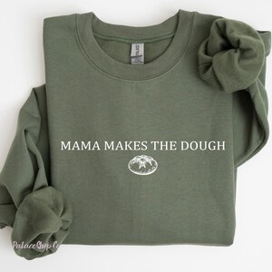 Può includere: Una felpa verde con la scritta "Mama Makes the Dough" e una piccola illustrazione di un dolce sul davanti.