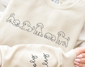 May include: A beige sweatshirt featuring a line of six black outline drawings of puppies in various poses. The sleeve cuff displays the names "Wooly" and "Bailey" in a cursive font. The sweatshirt is folded on a soft, light-colored surface.