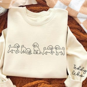 May include: Cream-colored sweatshirt with a crew neck and long sleeves. The front features a line of black line-art poodle illustrations. The right sleeve has the text "Teddy Echo" in a handwritten font. The sweatshirt is displayed on a textured brown surface.