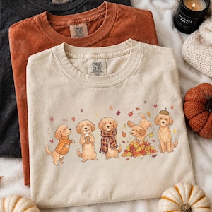 Peut inclure: Trois t-shirts à col rond en noir, orange brûlé et crème. Le t-shirt crème présente un motif de cinq chiens dorés vêtus d'accessoires d'automne, dont des pulls, des écharpes et un chapeau en forme de gland, entourés de feuilles tombantes.