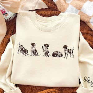Può includere: Felpa color crema con una fila di cinque illustrazioni di Pointer tedeschi in varie pose. Le maniche hanno polsini a coste e il nome "Sky" in corsivo sulla manica destra. La felpa è su una superficie marrone strutturata.