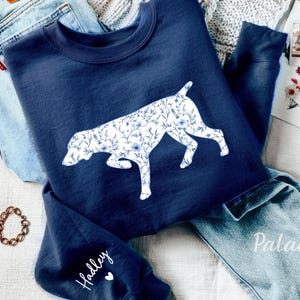 Puede incluir: Una sudadera azul marino con una silueta de perro blanca con estampado floral en la parte delantera. La sudadera tiene el nombre "Hadley" escrito en la manga.