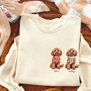 May include: Cream-colored sweatshirt featuring two brown dog illustrations with pink and blue bows. The names "Effie" and "Teddy" are printed below the dogs. The sweatshirt is displayed on a textured brown surface.