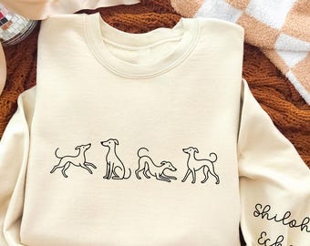 May include: Cream-colored sweatshirt featuring a line drawing of four dogs in various poses. The sleeve has the handwritten text "Shiloh Echo". The sweatshirt is displayed on a textured brown surface with a tan bow and a disco ball.