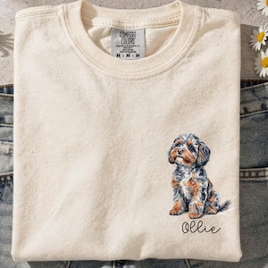 Puede incluir: Una camiseta de cuello redondo color crema con el gráfico de un perro pequeño y multicolor con pelaje marrón, negro y gris. Debajo del perro, el nombre Ollie está impreso en una tipografía cursiva negra. La camiseta tiene una etiqueta Comfort Colors.