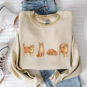 Könnte beinhalten: Beiges Sweatshirt mit goldenem Kettenausschnitt und einer Aquarellillustration von vier orangefarbenen Tabby-Katzen in verschiedenen Posen. Weiße High-Top-Sneaker und blaue Jeans sind ebenfalls im Bild.
