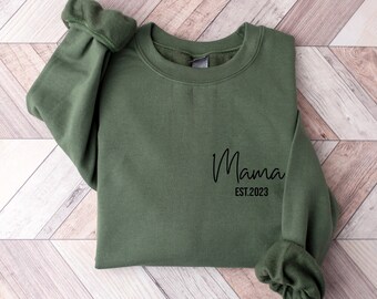 Könnte beinhalten: Ein grüner Pullover mit dem Aufdruck "Mama Est. 2023" auf der Vorderseite.