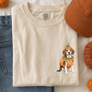 Könnte beinhalten: Ein cremefarbenes Kurzarm-T-Shirt mit einer Grafik eines Beagle-Hundes in einem Kürbiskostüm. Das T-Shirt hat ein Comfort Colors-Etikett am Halsausschnitt. Das Motiv befindet sich auf der unteren rechten Seite des Brustbereichs.