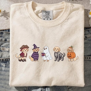 Puede incluir: Una camiseta de color crema con un gráfico de cinco perros de color marrón claro con varios disfraces de Halloween, incluyendo un pirata, una bruja, un fantasma, un esqueleto y una calabaza. La camiseta tiene una etiqueta Comfort Colors.