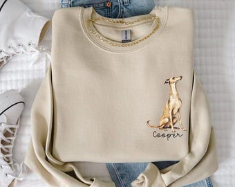 May include: Beige sweatshirt with a gold chain necklace and a graphic of a dog named "Cooper." The sweatshirt is paired with blue jeans and white high-top sneakers, all arranged on a white surface.