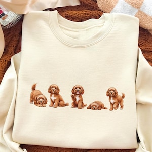 Könnte beinhalten: Cremefarbenes Sweatshirt mit einer Grafik von fünf braunen Welpen in verschiedenen Posen. Die Welpen sind realistisch dargestellt, mit detailliertem Fell und Ausdrücken. Das Sweatshirt hat einen klassischen Rundhalsausschnitt und lange Ärmel.