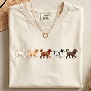 May include: A cream-colored crewneck t-shirt featuring a row of five small embroidered dogs in various colors including white, tan, reddish-brown, black and white spotted, and dark brown. The shirt has a Comfort Colors brand label at the neck.