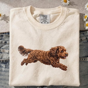 以下が含まれることがあります： クリーム色のComfort Colors Tシャツ。茶色の犬が走っているグラフィックが特徴です。犬は巻き毛で、舌を出しています。シャツはジーンズの上に折りたたまれ、周囲にはヒナギクが散らばっています。